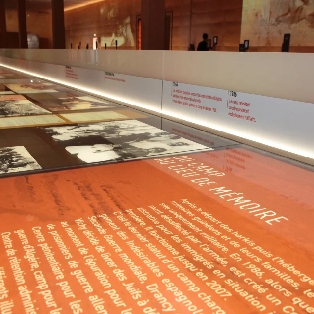Vitrine d’exposition au Mémorial du Camp de Rivesaltes présentant des textes, des photos d’archives et une frise chronologique retraçant l’histoire du camp.