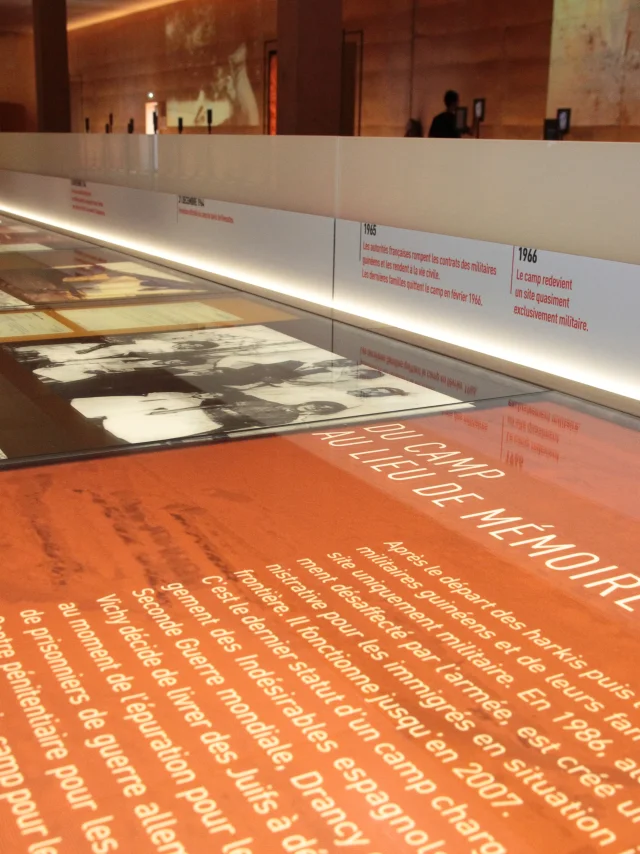 Vitrine d’exposition au Mémorial du Camp de Rivesaltes présentant des textes, des photos d’archives et une frise chronologique retraçant l’histoire du camp.