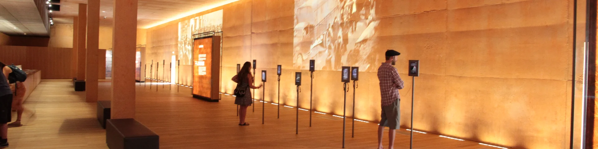 Visiteurs observant des écrans interactifs dans la grande salle d’exposition du Mémorial du Camp de Rivesaltes, aux murs ocre et à l’éclairage tamisé.