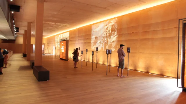 Visiteurs observant des écrans interactifs dans la grande salle d’exposition du Mémorial du Camp de Rivesaltes, aux murs ocre et à l’éclairage tamisé.