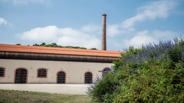 Bâtiment restauré de l’ancienne dynamiterie Nobel du site classé de Paulilles, avec sa haute cheminée en brique et des massifs fleuris au premier plan.