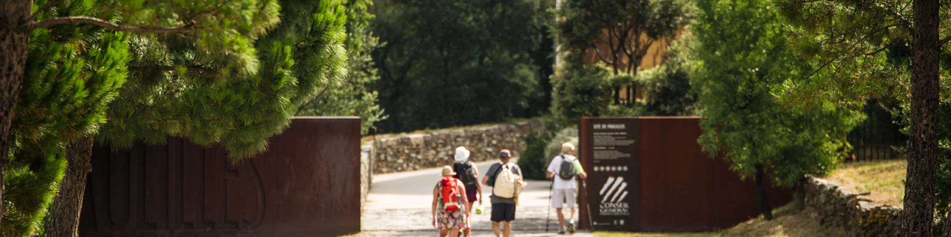 Entrée du site classé de Paulilles, à Port-Vendres, avec plusieurs promeneurs marchant sur une allée bordée de pins menant vers les bâtiments du site.