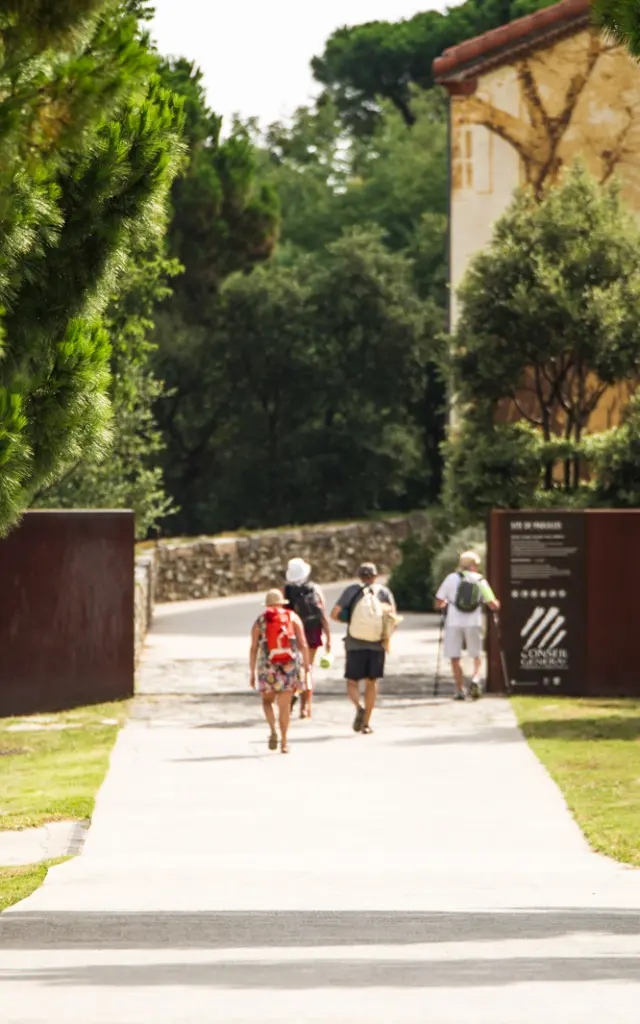 Entrée du site classé de Paulilles, à Port-Vendres, avec plusieurs promeneurs marchant sur une allée bordée de pins menant vers les bâtiments du site.