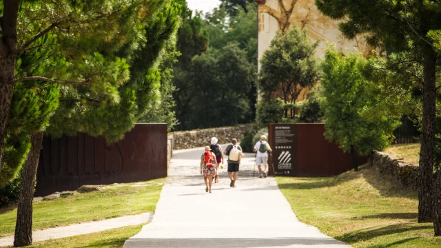 Entrée du site classé de Paulilles, à Port-Vendres, avec plusieurs promeneurs marchant sur une allée bordée de pins menant vers les bâtiments du site.