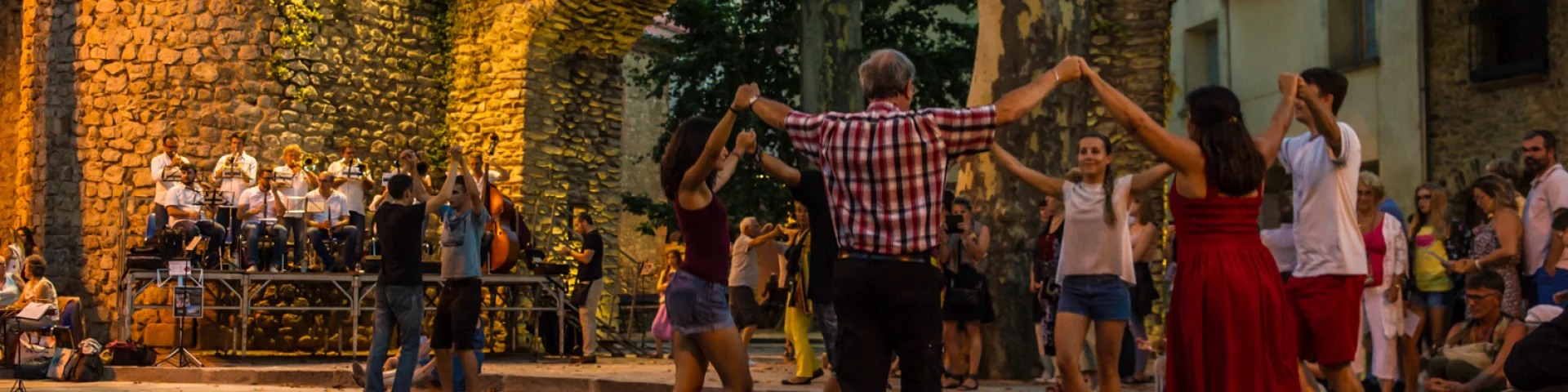 Groupe de personnes dansant la sardane en cercle sur une place de Céret, devant un orchestre de cobla et des arcades en pierre décorées de drapeaux catalans.