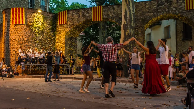 Groupe de personnes dansant la sardane en cercle sur une place de Céret, devant un orchestre de cobla et des arcades en pierre décorées de drapeaux catalans.