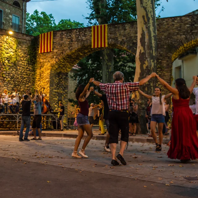 Groupe de personnes dansant la sardane en cercle sur une place de Céret, devant un orchestre de cobla et des arcades en pierre décorées de drapeaux catalans.