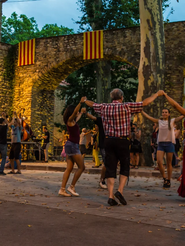 Groupe de personnes dansant la sardane en cercle sur une place de Céret, devant un orchestre de cobla et des arcades en pierre décorées de drapeaux catalans.