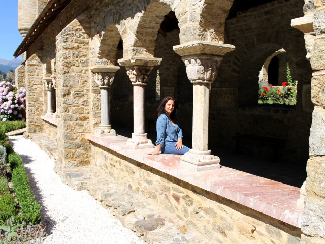 Femme assise dans le cloître en pierre de l’abbaye Saint-Martin du Canigou, entourée d’arcades romanes sculptées et d’un jardin fleuri.
