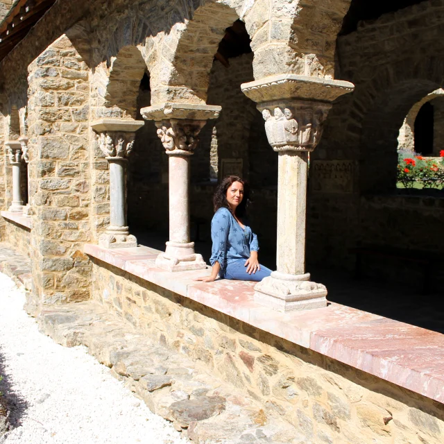 Femme assise dans le cloître en pierre de l’abbaye Saint-Martin du Canigou, entourée d’arcades romanes sculptées et d’un jardin fleuri.