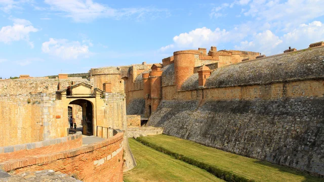Entrée et remparts en pierre et brique de la forteresse de Salses, sous un ciel bleu partiellement nuageux.