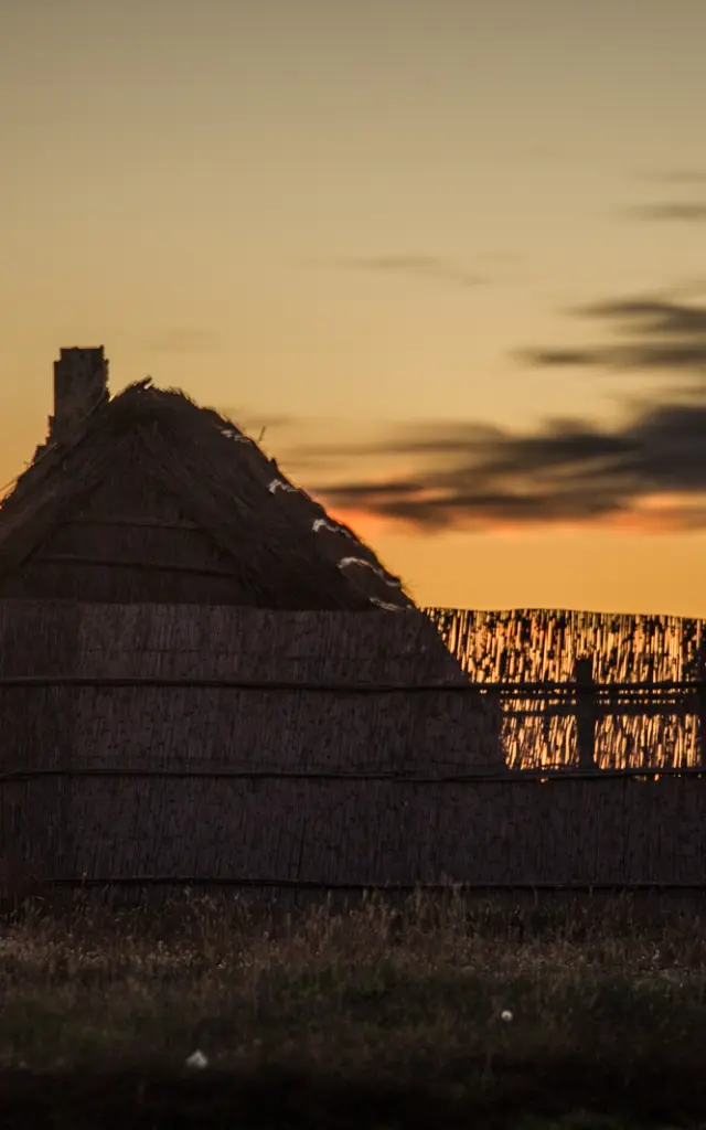 Cabane traditionnelle en roseaux silhouettée au coucher du soleil, sur fond de ciel doré et orangé.