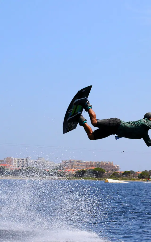 Pratiquant de wakeboard effectuant une figure aérienne sur un plan d’eau, tiré par un câble, sous un ciel bleu à proximité du littoral urbain.