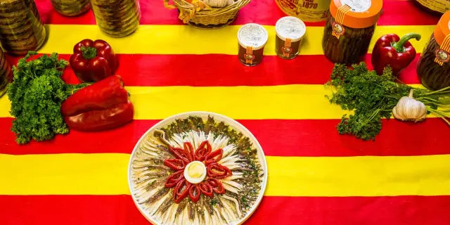 Assiette d’anchois de Collioure entourée de poivrons rouges et de persil, présentée sur une nappe aux couleurs catalanes rouge et jaune.