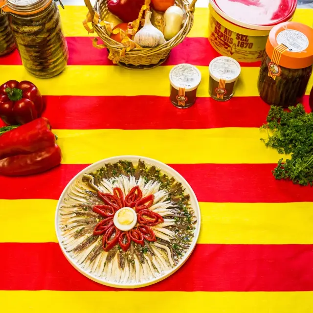 Assiette d’anchois de Collioure entourée de poivrons rouges et de persil, présentée sur une nappe aux couleurs catalanes rouge et jaune.