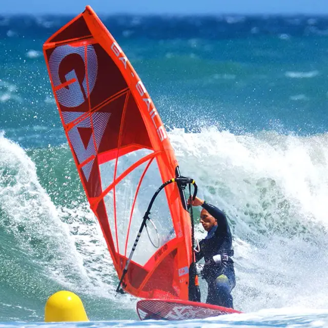 Planche à voile rouge affrontant une grande vague sur la mer Méditerranée, sous un ciel bleu éclatant.