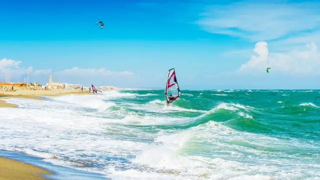 Planche à voile et kitesurf sur une plage méditerranéenne balayée par le vent, sous un ciel bleu lumineux.