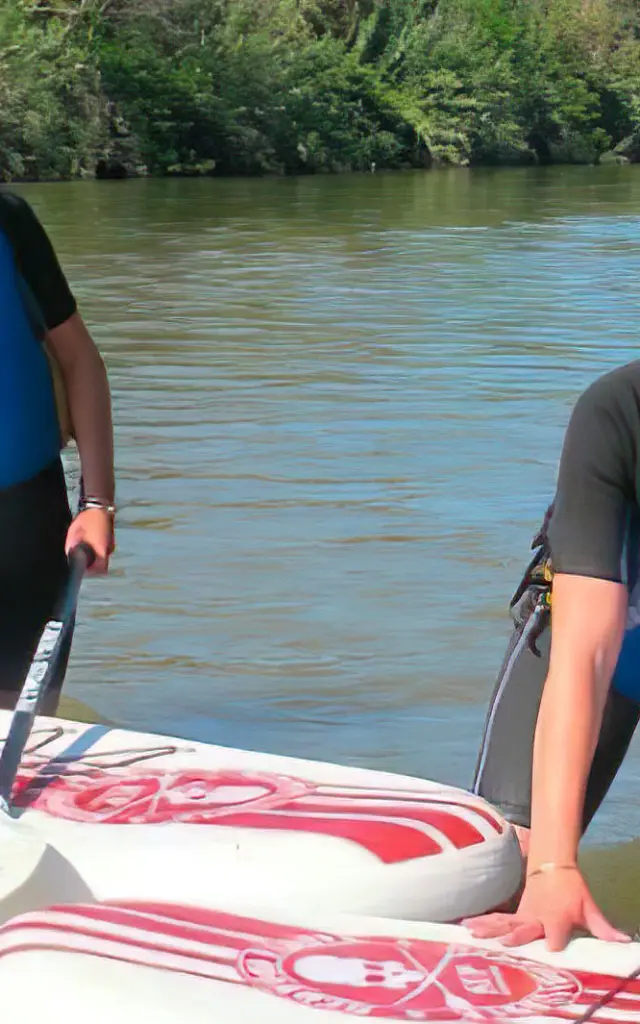 Deux personnes en combinaison et gilet de sauvetage pratiquent le stand up paddle sur une rivière bordée d’arbres.