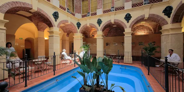 Galerie thermale voûtée en briques et pierres avec bassin d’eau bleue et plantes, fréquentée par des curistes en peignoir.