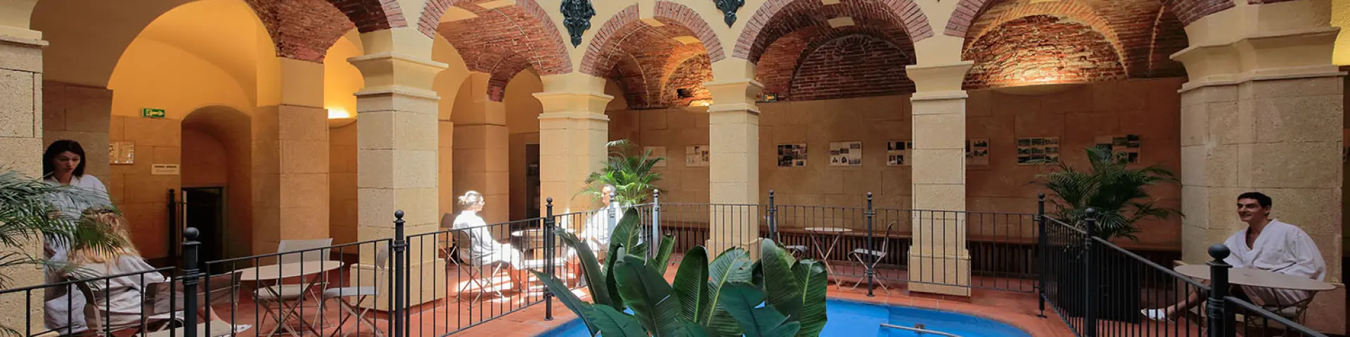 Galerie thermale voûtée en briques et pierres avec bassin d’eau bleue et plantes, fréquentée par des curistes en peignoir.