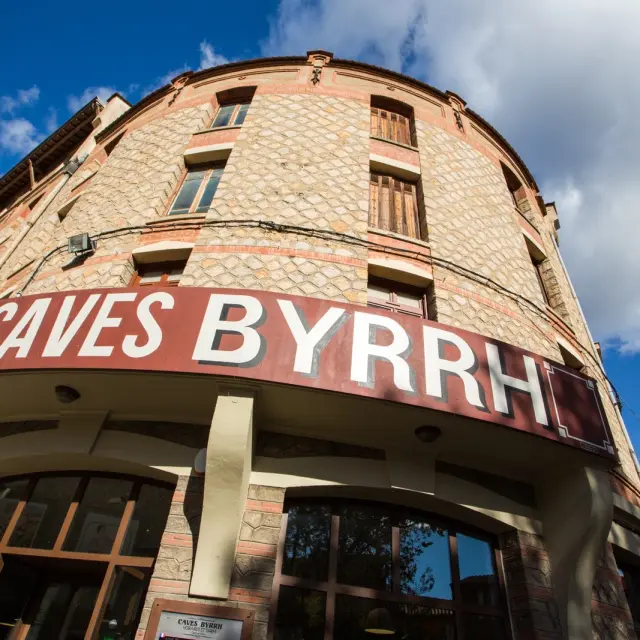 Façade en pierre des Caves Byrrh à Thuir, surmontée d’une grande enseigne rouge et blanche sous un ciel bleu parsemé de nuages.
