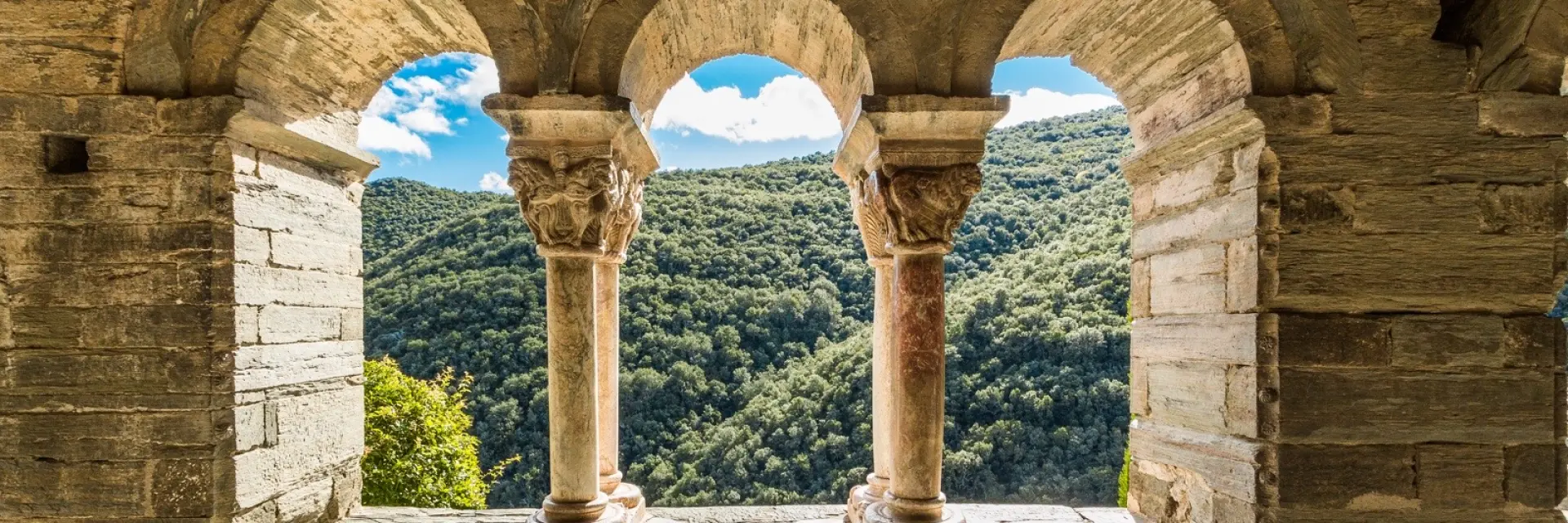 Vue à travers les arcades d’un cloître roman en pierre, ouvrant sur les collines verdoyantes d’une vallée ensoleillée.
