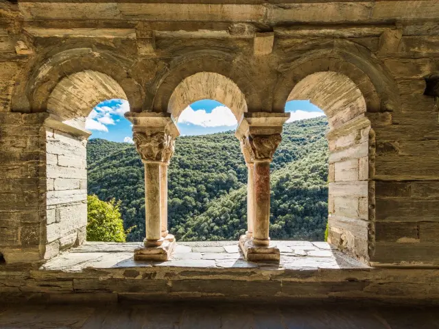 Vue à travers les arcades d’un cloître roman en pierre, ouvrant sur les collines verdoyantes d’une vallée ensoleillée.