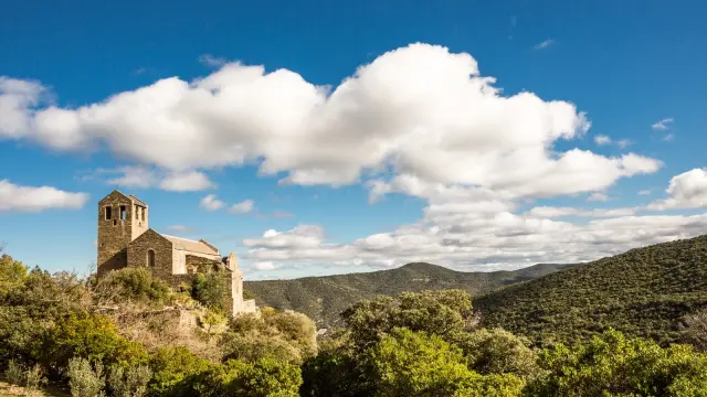Église romane en pierre perchée sur une colline verdoyante, entourée de garrigue et de montagnes sous un ciel bleu parsemé de nuages blancs.