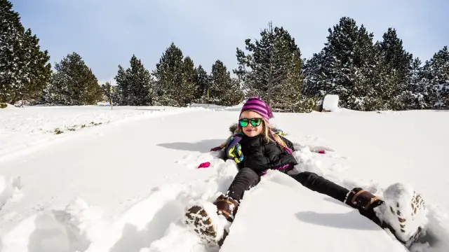 Enfant souriant allongé dans la neige, portant bonnet violet et lunettes de soleil, entouré d’arbres enneigés sous un ciel clair.