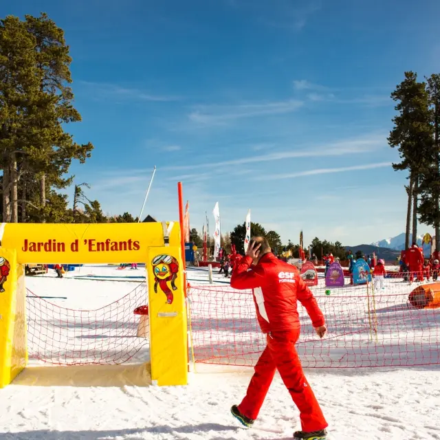 Moniteur de ski en combinaison rouge passant devant l’entrée du Jardin d’Enfants de l’ESF à Font-Romeu, sous un ciel bleu et face aux montagnes enneigées.