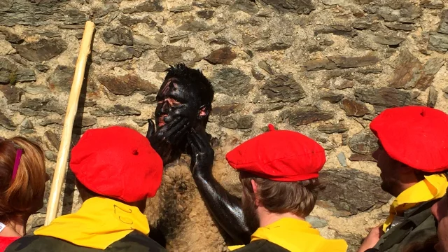 Participant de la fête de l’Ours à Prats-de-Mollo-la-Preste, le visage couvert de noir, entouré d’hommes coiffés de bérets rouges et vêtus de foulards jaunes, devant un mur de pierre.