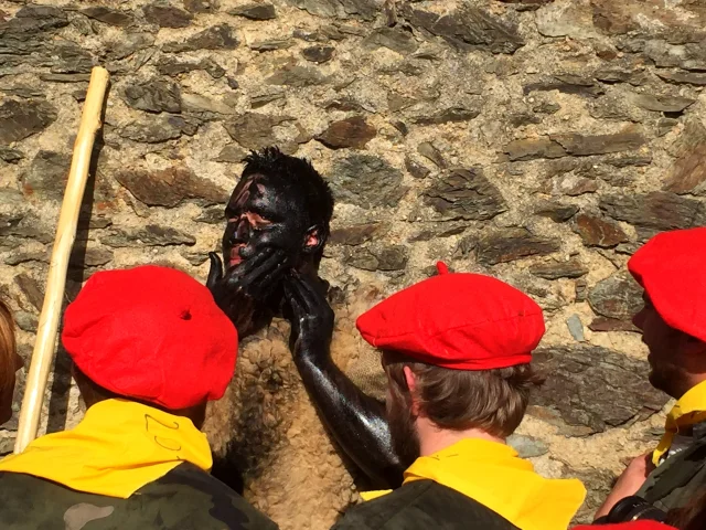 Participant de la fête de l’Ours à Prats-de-Mollo-la-Preste, le visage couvert de noir, entouré d’hommes coiffés de bérets rouges et vêtus de foulards jaunes, devant un mur de pierre.