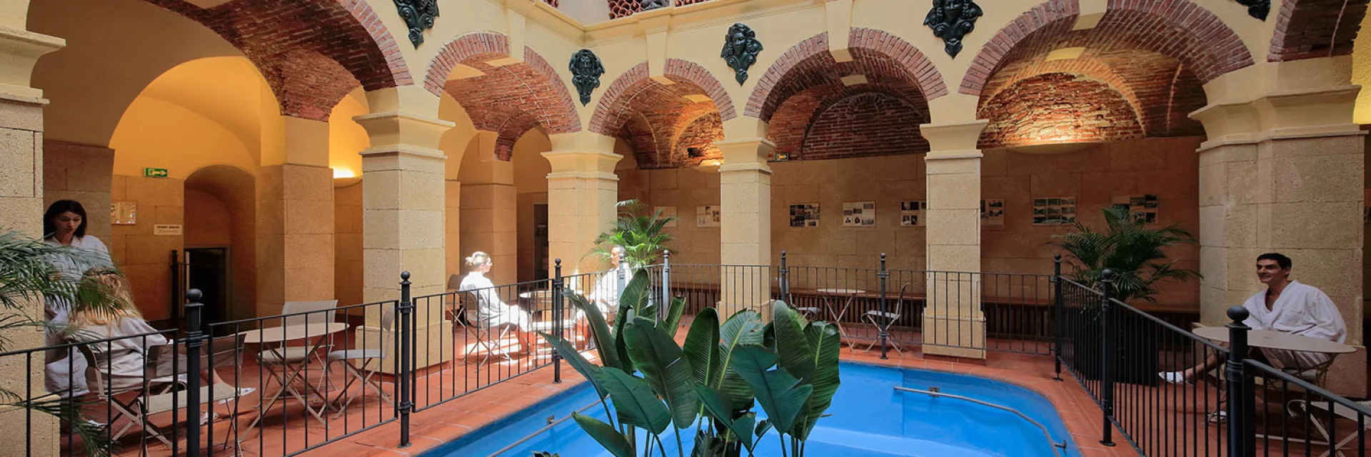 Galerie thermale voûtée en briques et pierres avec bassin d’eau bleue et plantes, fréquentée par des curistes en peignoir.