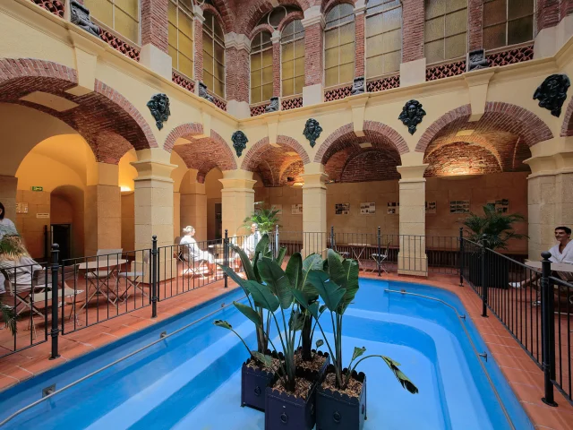 Galerie thermale voûtée en briques et pierres avec bassin d’eau bleue et plantes, fréquentée par des curistes en peignoir.