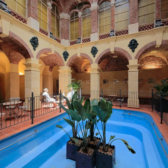 Galerie thermale voûtée en briques et pierres avec bassin d’eau bleue et plantes, fréquentée par des curistes en peignoir.