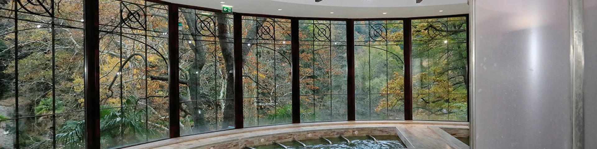 Deux personnes se détendent dans une piscine thermale intérieure entourée de grandes baies vitrées ouvrant sur la forêt.