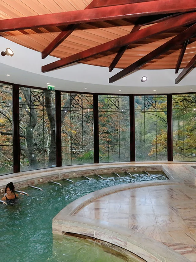 Deux personnes se détendent dans une piscine thermale intérieure entourée de grandes baies vitrées ouvrant sur la forêt.