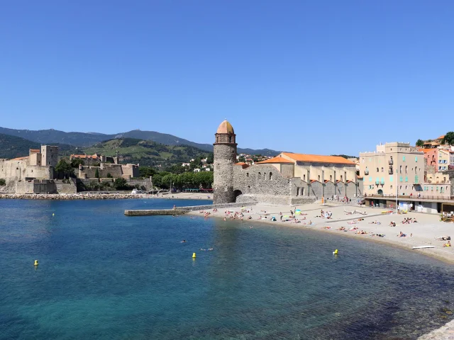 Vue sur la plage de Collioure avec son clocher emblématique, les façades colorées du village et le Château Royal en arrière-plan, sous un ciel bleu.