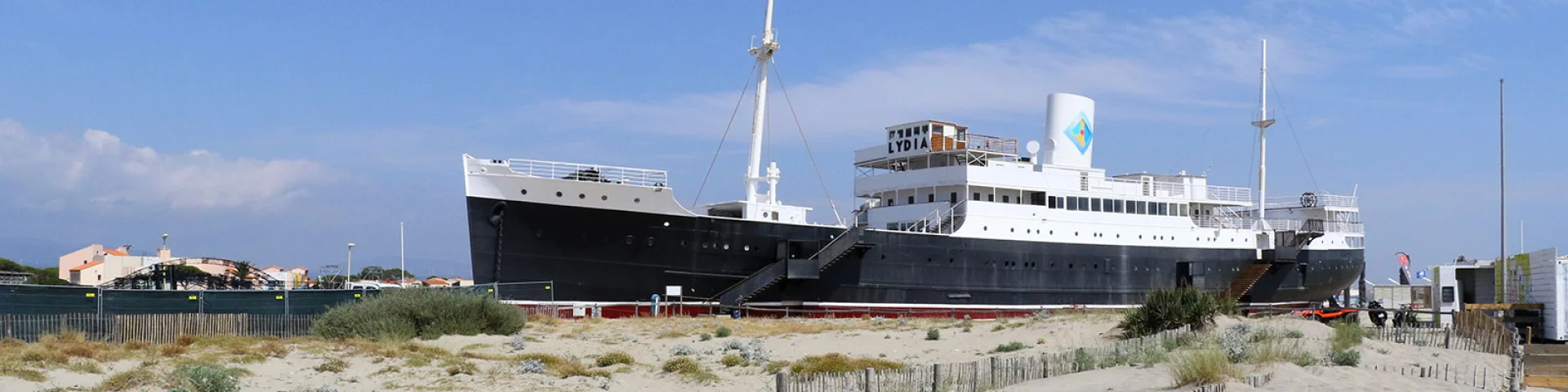Le paquebot Lydia échoué sur la plage du Barcarès, vu depuis les dunes de sable et la végétation littorale, sous un ciel bleu dégagé.