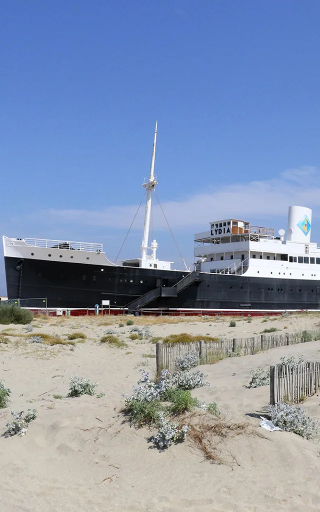 Le paquebot Lydia échoué sur la plage du Barcarès, vu depuis les dunes de sable et la végétation littorale, sous un ciel bleu dégagé.