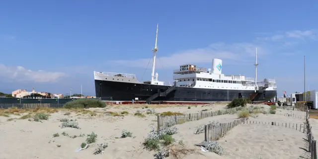 Le paquebot Lydia échoué sur la plage du Barcarès, vu depuis les dunes de sable et la végétation littorale, sous un ciel bleu dégagé.
