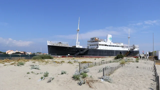 Le paquebot Lydia échoué sur la plage du Barcarès, vu depuis les dunes de sable et la végétation littorale, sous un ciel bleu dégagé.