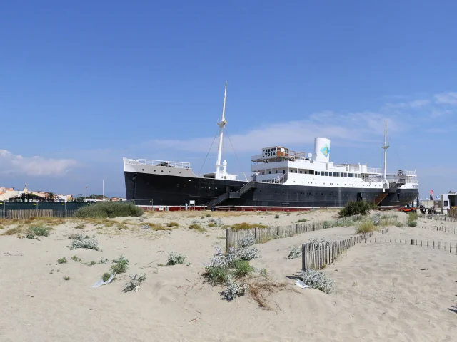 Le paquebot Lydia échoué sur la plage du Barcarès, vu depuis les dunes de sable et la végétation littorale, sous un ciel bleu dégagé.