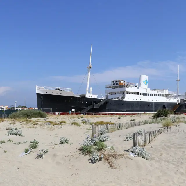 Le paquebot Lydia échoué sur la plage du Barcarès, vu depuis les dunes de sable et la végétation littorale, sous un ciel bleu dégagé.