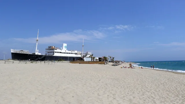 Le paquebot Lydia échoué sur la plage du Barcarès, sous un grand ciel bleu, avec quelques baigneurs profitant du bord de mer.