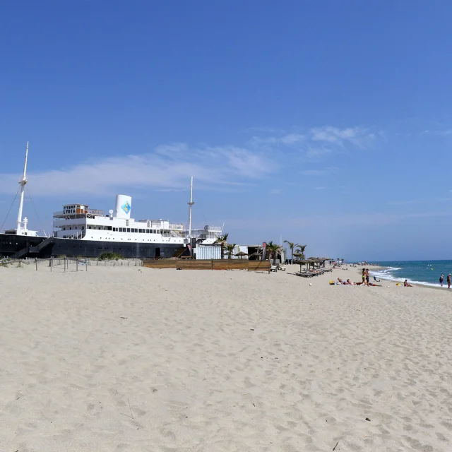 Le paquebot Lydia échoué sur la plage du Barcarès, sous un grand ciel bleu, avec quelques baigneurs profitant du bord de mer.