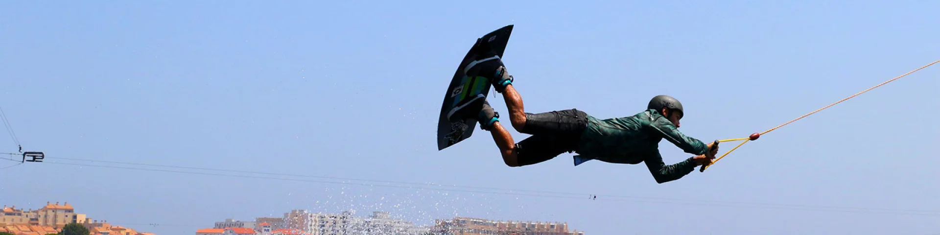 Pratiquant de wakeboard effectuant une figure aérienne sur un plan d’eau, tiré par un câble, sous un ciel bleu à proximité du littoral urbain.