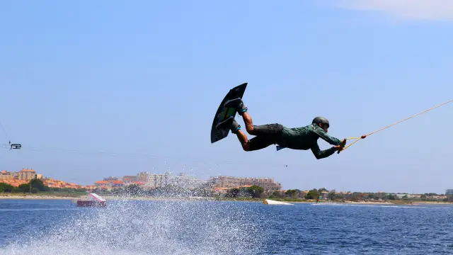 Pratiquant de wakeboard effectuant une figure aérienne sur un plan d’eau, tiré par un câble, sous un ciel bleu à proximité du littoral urbain.