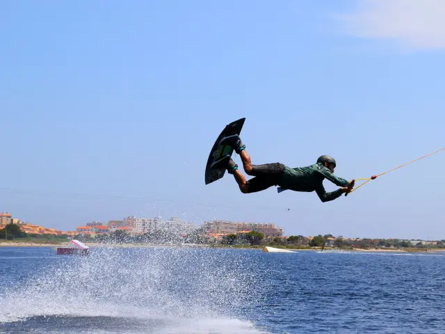 Pratiquant de wakeboard effectuant une figure aérienne sur un plan d’eau, tiré par un câble, sous un ciel bleu à proximité du littoral urbain.