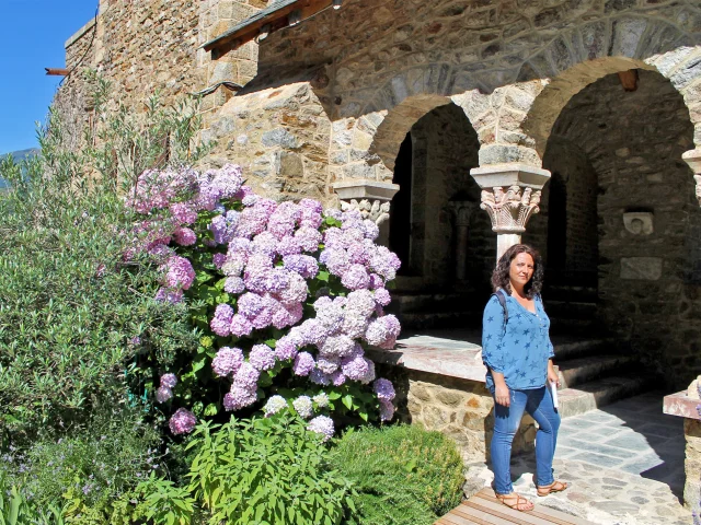 Personne debout à l’entrée du cloître de l’abbaye Saint-Martin du Canigou, entourée de fleurs et de murs en pierre.
